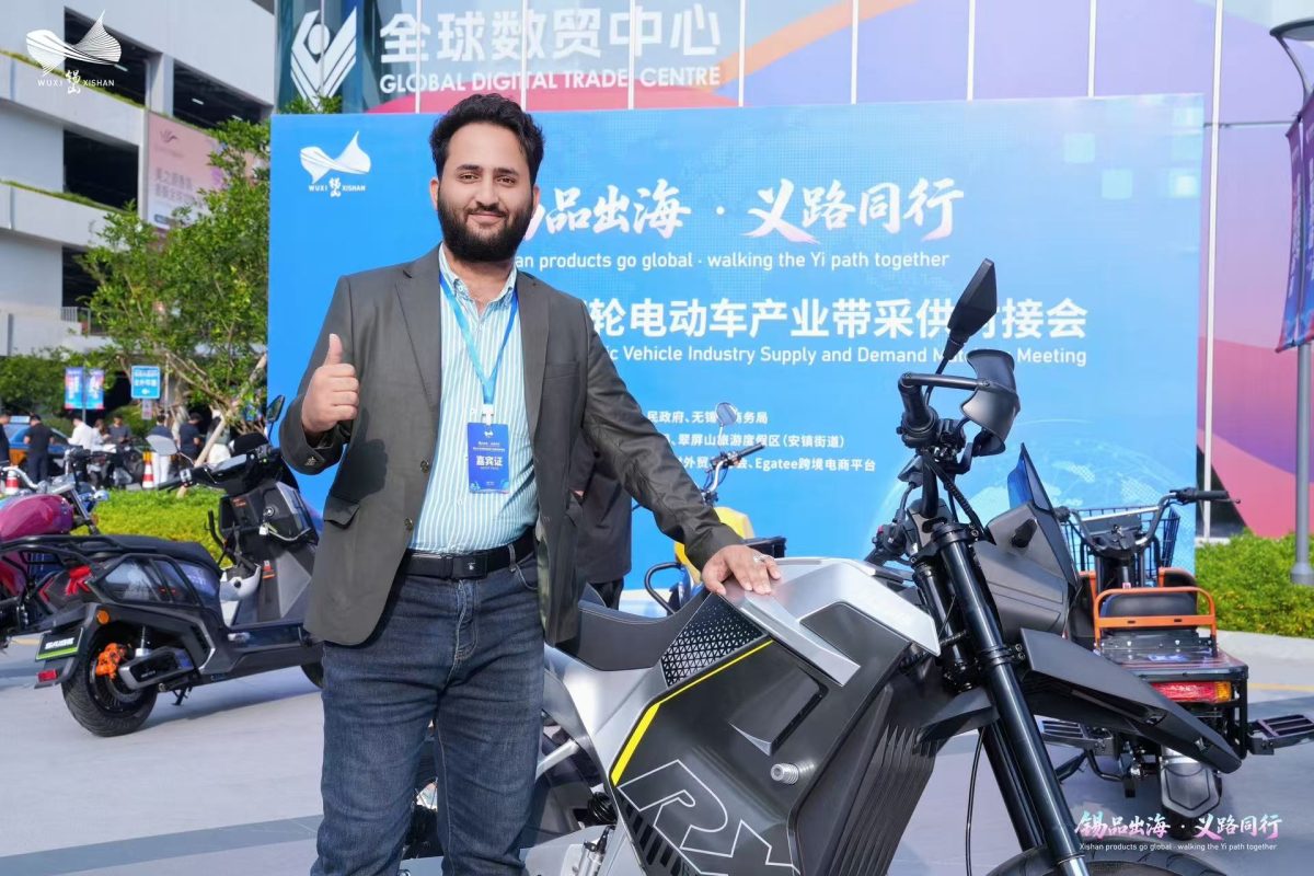 International buyer gives a thumbs-up beside a silver electric motorcycle at the product display.