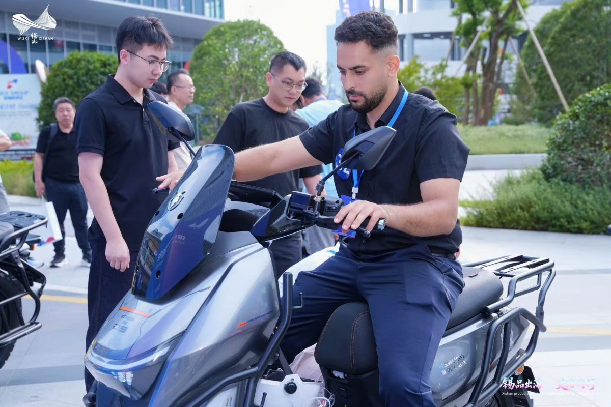 Overseas buyer tests the controls of a gray electric scooter while supplier representatives observe.